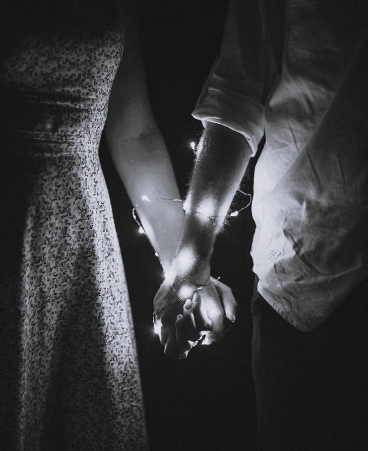 A couple holds hands in soft light with string lights wrapped around their wrists. Power in a relationship can be both connection and control. A relationship therapist in Delray Beach, FL, helps partners rediscover healthy emotional balance and connection.