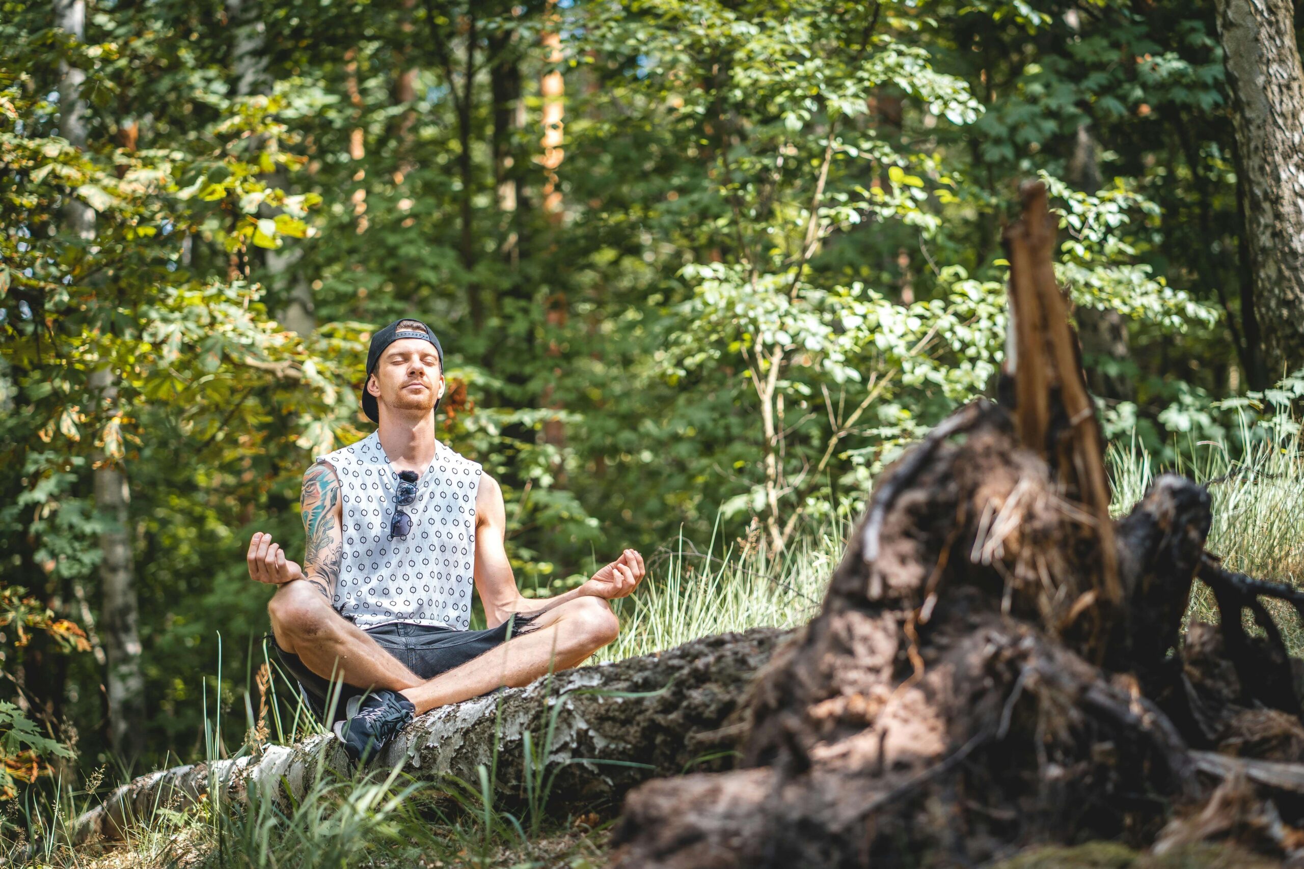 Man meditating | meditation therapy for anxiety delray beach | anxiety therapy | mindfulness therapy | Delray Beach, Florida | Juno Beach | Loxahatchee | Village by the Sea | Pineapple Grove | SOFA | Lake Ida A man in a cap meditates in lotus position on a fallen log in a lush forest. Meditation helps calm the overactive mind and reduce the grip anxiety has on daily life. Anxiety therapy in Delray Beach, FL, can help you find your path to peace.