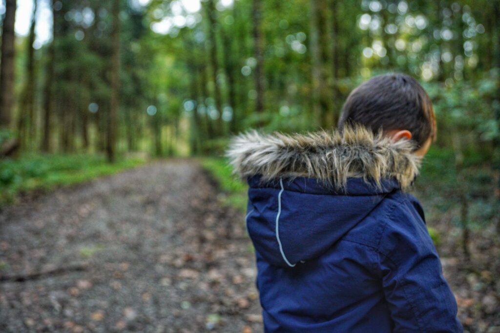 Child wearing a navy jacket with a fur-trimmed hood, viewed from behind while standing along a wooded trail.