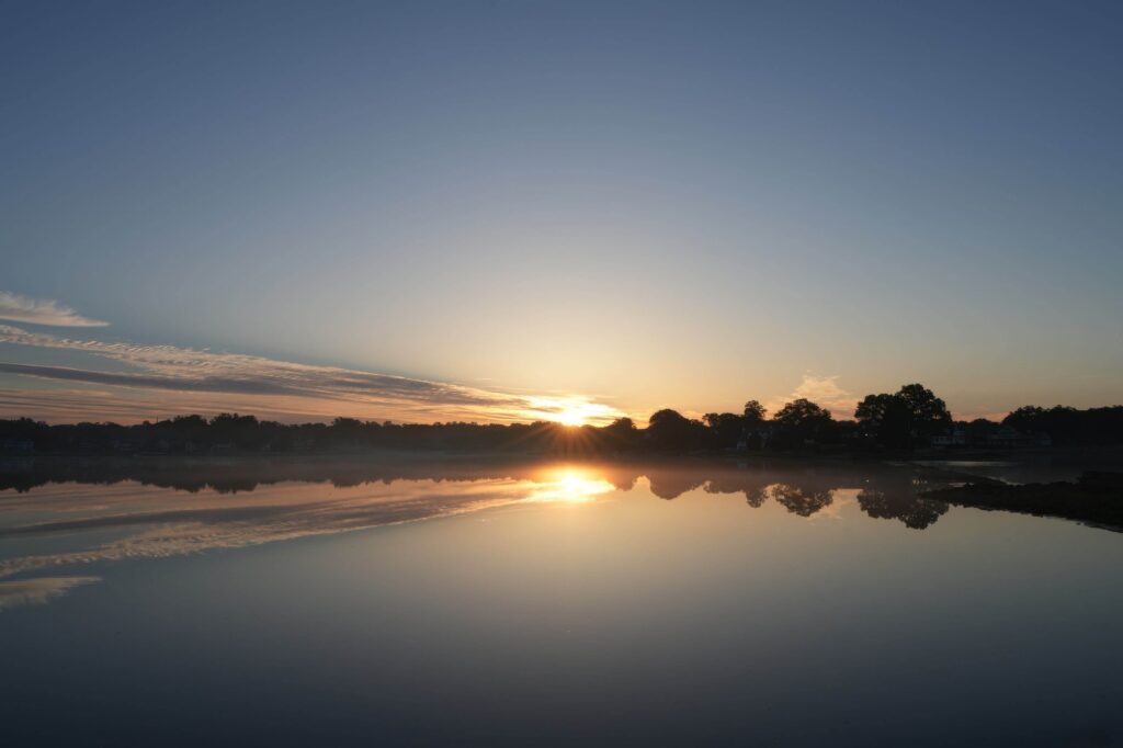 Sun rising over a calm lake, casting golden light across the water and reflecting trees along the distant shoreline.