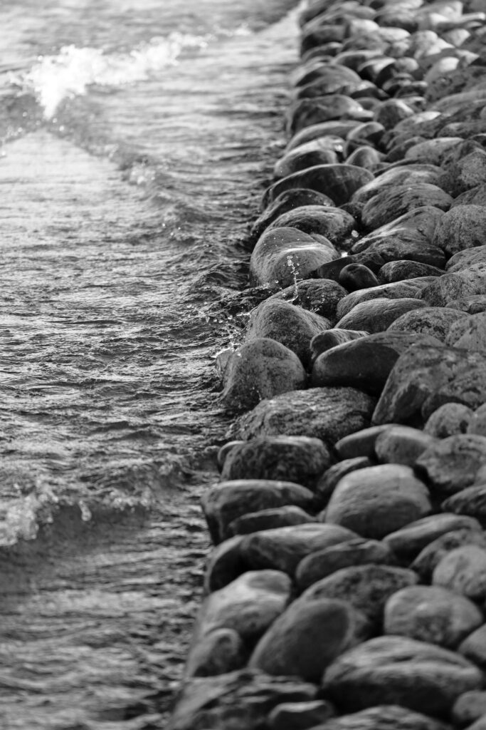 Black-and-white close-up of gentle waves washing against smooth, rounded rocks along a shoreline.