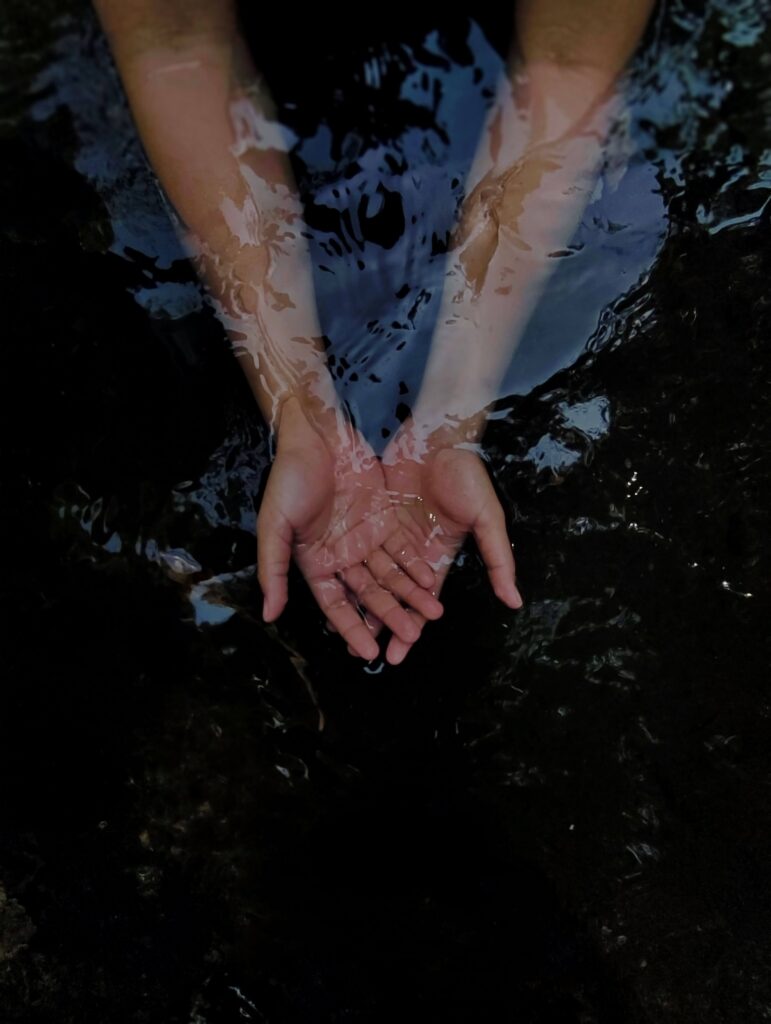 Close-up of two open hands resting just beneath the surface of dark water, surrounded by gentle ripples and reflections.