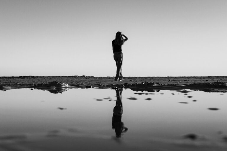 Black-and-white image of a woman standing alone near the water, with her silhouette reflected in the still surface below.
